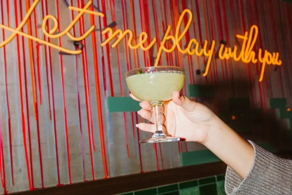 A hand holding a premium craft cocktail during a Happy Hour Chicago event at AceBounce, featuring an energetic neon sign background reading "let me play with you."