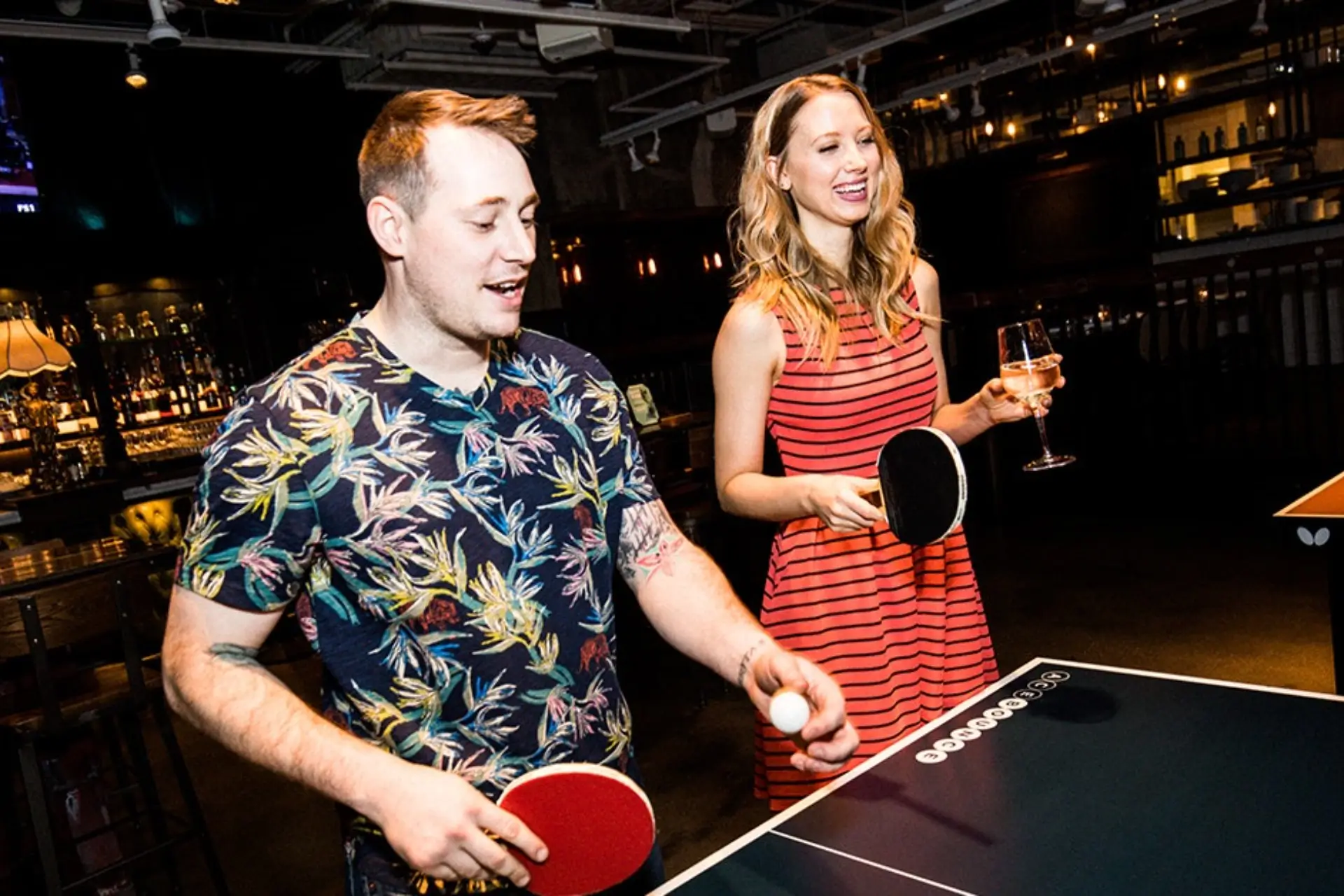 A man and woman enjoying drinks and playing ping pong at AceBounce while watching sports on TV, demonstrating social viewing and interactive group games during a Chicago sports outing.